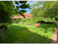 Grüne Gartenanlage mit Rasenfläche, umgeben von Bäumen und Sträuchern. Mehrere Sitzgruppen aus Holzmöbeln und ein roter Stuhl stehen auf der Wiese, im Hintergrund sind ein Gebäude mit roten Ziegeldächern und Hecken zu sehen.