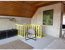 Zimmer in einem Dachgeschoss mit Holzdeckenverkleidung, beigem Teppichboden und einer gelben Treppenbrüstung. An der Wand hängt ein Bild mit Waldmotiv, daneben steht ein schwarzer Kleiderschrank mit offenen Kleiderbügeln und ein moderner Stuhl mit Kissen.