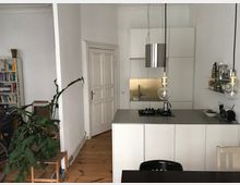 Ein minimalistisch eingerichteter Wohn- und Küchenbereich mit weißen Wänden und Holzdielenboden. Im Vordergrund befindet sich eine moderne Kücheninsel mit Hängelampen, dahinter eine integrierte Küche mit weißen Schränken und einer goldfarbenen Rückwand; links ein Regal mit Büchern und eine große Zimmerpflanze.