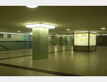 Unterirdischer U-Bahn-Bereich mit gelb-grünen Fliesenwänden, zentralen beleuchteten Säulen und einem rechteckigen Fahrplan-Leuchtkasten. Im Hintergrund befinden sich Treppen und Geländer, die zu verschiedenen Ausgängen führen.