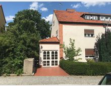 Zweistöckiges Wohnhaus mit hell verputzter Fassade, rotem Ziegeldach und einem kleinen Anbau mit weißen Türen im Eingangsbereich. Vor dem Haus befinden sich ein gepflasterter Gehweg, grüne Hecken und Bäume.