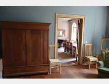 Ein stilvoll eingerichteter Innenraum mit einem antiken Holzschrank und zwei Holzstühlen im Vordergrund. Im Hintergrund ist ein Esszimmer mit Holztisch und Stühlen sowie großen, bogenförmigen Fenstern zu sehen.