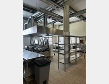 Große, industriell ausgestattete Küche mit freistehenden Metallarbeitsflächen, Regalen und Spülstationen. Sichtbare Lüftungsrohre verlaufen unter der Decke, während der Boden mit beigen Fliesen ausgelegt ist.