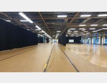 Großer Veranstaltungsraum mit gefliesten Böden, schwarzen Vorhängen als Raumteiler und einer offenen Deckenkonstruktion mit sichtbaren Leuchten. An den Seiten befinden sich Fensterfronten, die Tageslicht hereinlassen.