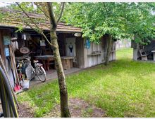 Holzschuppen mit Satteldach in einer ländlichen Umgebung, umgeben von grüner Wiese und Bäumen. Vor dem Schuppen stehen verschiedene Gartengeräte, ein Fahrrad und rustikale Möbel; der Innenbereich ist teilweise sichtbar.