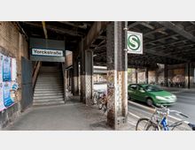 Ansicht eines Bahnhofsbereichs an der Yorckstraße in Berlin unter einer historischen Eisenbahnbrücke mit genieteten Stahlträgern. Links führen Treppen zu einer S-Bahn-Station, während Fahrräder an Pfeilern geparkt sind und Autos die angrenzende Straße passieren.