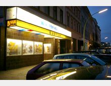 Straßenansicht eines kleinen Kinos mit dem Namen "Babylon", erkennbar am beleuchteten gelben Vordach mit Filmtiteln und Schaukastenfenstern mit Filmplakaten an der mit dunklen Fliesen verkleideten Fassade. Mehrere parkende Autos stehen auf der Straße, und im Hintergrund sind Wohnhäuser sowie Straßenlaternen sichtbar.