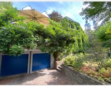 Ein zweistöckiges Wohnhaus, das vollständig mit grünem Efeu bewachsen ist. Vor dem Gebäude befinden sich zwei blaue Garagentore, eine steinerne Gartenmauer mit Pflanzen sowie ein gepflasterter Weg.