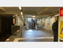 U-Bahn-Station mit Treppenaufgang zum Ausgang, flankiert von weißen, gefliesten Wänden und beleuchtet durch Deckenlampen. Links ist ein Gleisbereich erkennbar, rechts befinden sich Werbeplakate und ein Fahrscheinautomat.