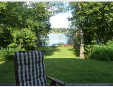 Ein Garten mit Rasenfläche, umgeben von Bäumen und Büschen, führt zu einem See im Hintergrund. Im Vordergrund steht ein gepolsterter Gartenstuhl, während am Seeufer eine Holzbank und ein Baumhaus zu erkennen sind; auf dem See fährt ein kleines Boot.