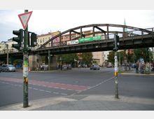 Straßenkreuzung in einer Stadt mit einer erhöhten Eisenbahnbrücke aus Metall im Hintergrund, umgeben von Wohn- und Geschäftshäusern. Im Vordergrund sind Ampeln, ein Zebrastreifen und Straßenschilder sichtbar, mit Personen, Autos und Fahrrädern im Straßenbereich.