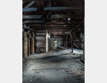 Innenansicht eines alten, verlassenen Dachbodens mit sichtbaren Holzbalken, beschädigtem Boden und bröckelnden Wänden. In der Mitte führt eine hölzerne Leiter zu einem höheren Bereich, und eine offene Tür im Hintergrund lässt Licht herein.