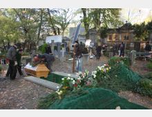 Ein Friedhof mit einem offenen Grab, in das ein Holzsarg mit Blumenschmuck abgesenkt wird. Umgeben sind das Grab und umliegende Gräber von Bäumen, gefallenen Blättern und alten Grabsteinen, während Filmcrew und Ausrüstung auf der Szene zu sehen sind.