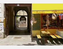 Eine schmale Gasse mit einem Torbogen führt zu einem Innenhof, daneben ein Geschäft mit gelber Fassade und der Aufschrift „Knofi“. Vor dem Geschäft stehen Holzbänke und -tische, dekoriert mit Knoblauchzöpfen und einer Fahne, während eine handgeschriebene Tafel Preise anzeigt.