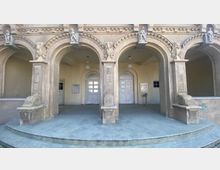 Historische Fassade eines Gebäudes mit drei großen Bögen, verziert mit Statuen und Reliefs. Die Bögen führen zu weißen Türen, und die Architektur zeigt klassische Details wie Säulen und geschwungene Verzierungen.