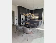 Moderne Küche mit dunklen Einbauschränken, einer Kücheninsel und integriertem Backofen. Im Vordergrund befindet sich ein Esstisch mit schwarzer Glasplatte, umgeben von grauen Polsterstühlen, und darüber hängen mehrere goldene Pendelleuchten.