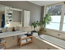 Modernes Badezimmer mit großem Waschbecken mit Doppelarmaturen, Spiegeln und weißen Schränken. Eine Badewanne befindet sich unter einem großen Fenster mit satiniertem Glas, umgeben von Topfpflanzen; darunter stehen Holzmöbel mit gestapelten Handtüchern.