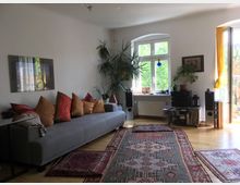 Wohnzimmer mit hellem Holzboden, einer grauen Couch mit farbigen Kissen und mehreren orientalischen Teppichen. Große Fenster mit gelben Vorhängen und ein Balkonausgang lassen viel Tageslicht herein; Zimmerpflanzen und eine Stereoanlage auf einem Regal ergänzen die Einrichtung.