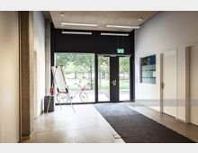 Moderner Eingangsbereich mit großen Glastüren, die zu einem Außenbereich mit Bäumen führen. Im Raum befinden sich ein Flipchart, ein Mülleimer und ein schwarzer Teppich auf einem Betonboden; draußen ist ein abgestelltes Fahrrad sichtbar.