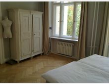 Helles Schlafzimmer mit Holzparkettboden, einem weißen, antik wirkenden Kleiderschrank und einer Schaufensterpuppe. Große Fenster mit beigem Vorhang bieten Ausblick auf Bäume, darunter ein Heizkörper.