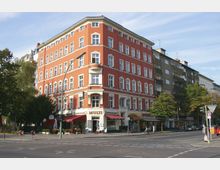 Eckgebäude mit roter Fassade und weißen Fensterrahmen in einem urbanen Wohnviertel, im Erdgeschoss befinden sich Geschäfte und Cafés mit Markisen. Im Vordergrund verläuft eine Straßenkreuzung mit Verkehrsschildern und Gehwegen, umgeben von Bäumen.