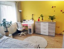 Ein helles Schlafzimmer mit einer gelben Wand, dekoriert mit Schmetterlingsaufklebern. Der Raum enthält ein Bett, einen Schreibtisch mit Drehstuhl, eine Kommode mit Zimmerpflanzen darauf, einen runden hellen Teppich und ein großes Fenster mit weißen Vorhängen.