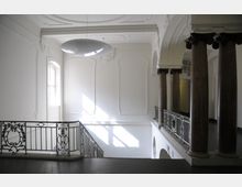 Innenraum eines historischen Gebäudes mit hohen Decken und verzierten Wandpaneelen in Weiß. Eine Treppe mit kunstvollem Metallgeländer und mehrere massive Säulen mit korinthischen Kapitellen prägen den Raum, der von natürlichem Licht durch ein Fenster erhellt wird.