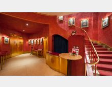 Eingangsbereich eines Theaters mit roten Wänden und vergoldeten Türen; an den Wänden hängen gerahmte Bilder mit Beleuchtung, rechts führt eine geschwungene Treppe mit rotem Teppich hinauf, und im Vordergrund stehen Tische mit floralem Dekor.