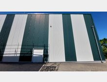 Außenfassade eines Industriegebäudes mit vertikalen Paneelen in dunkelgrüner und weißer Streifenoptik, kleiner weißer Tür am unteren Rand und einem gepflasterten Vorplatz. Ein blauer Himmel und etwas Vegetation sind am rechten Bildrand erkennbar.