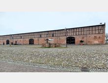 Großer Backsteinbau mit Fachwerkelementen und breiten Holztoren, umgeben von einem weitläufigen, gepflasterten Platz. Im Vordergrund steht ein kleiner gemauerter Brunnen mit Metallabdeckung.