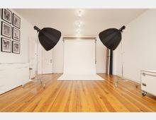 Ein Fotostudio mit einem weißen Hintergrund, Holzboden und zwei großen schwarzen Studioleuchten auf Ständern. An der linken Wand hängen gerahmte Schwarz-Weiß-Fotografien, und rechts stehen aufgerollte farbige Hintergründe.