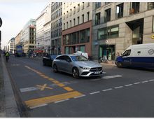 Straßenszene in einer Stadt mit mehrstöckigen modernen Gebäuden und Schaufenstern. Auf der Straße fahren Autos, darunter ein silberner Mercedes, sowie ein Lieferwagen; Radwege und Markierungen sind sichtbar, Fußgänger und Fahrräder befinden sich am Straßenrand.