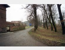 Ein unbefestigter Weg führt zwischen einer Backsteinmauer auf der linken Seite und einer von Bäumen gesäumten Grünfläche auf der rechten Seite entlang. Im Hintergrund sind weitere Backsteingebäude sichtbar, und ein blaues Verkehrsschild steht am Rand des Weges.