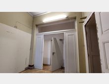 Ein einfacher Innenraum mit beige gestrichenen Wänden und abgenutztem Zustand, zeigt eine geöffnete weiße Tür mit schwarzem Türgriff in einem Türrahmen mit weißen Zierleisten. Über der Tür befindet sich eine Leuchtstoffröhre.