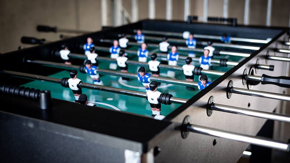 Foosball table indoors with a green playing surface and rows of miniature players in blue and white uniforms mounted on metal rods. The black table frame and handles are visible along the sides in a softly lit room.
