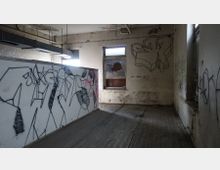 Verlassener, heruntergekommener Raum mit abgenutztem Holzboden, Graffiti an den Wänden und einem zerbrochenen Fenster, durch das gedämpftes Licht einfällt. Decke und Wände zeigen Spuren von Verfall und Schmutz.