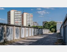 Mehrstöckige Wohnblocks mit Balkonen im Hintergrund, davor eine schmale, gepflasterte Gasse mit langen Reihen von kleinen Garagen mit Metalltoren, umgeben von vereinzelten Bäumen und Büschen.