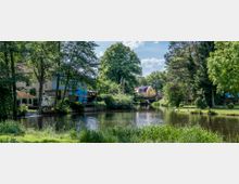 Landschaft mit einem ruhigen Fluss, umgeben von Bäumen und Wiesen. Im Hintergrund stehen farbige Häuser, darunter ein gelbes und ein blaues, sowie eine Brücke über den Fluss.
