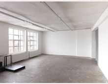 Ein leerer Raum mit kahlen weißen Wänden, großen Sprossenfenstern, grauem Betonboden und sichtbaren Heizkörpern unter den Fenstern. Von der Decke hängen lange, schlichte Lichtschienen, und links im Bild befinden sich dunkle, minimalistische Podeste.