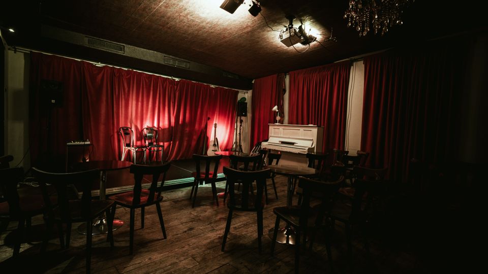 Small performance room with a low stage backed by deep red curtains, two chairs under stage lights, and musical instruments nearby. The space has wooden plank flooring, café-style tables and chairs facing the stage, a white upright piano, and ceiling-mounted lights with a chandelier.