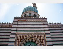 Nahaufnahme der Fassade einer Moschee mit gestreiftem Mauerwerk aus roten, weißen und grünen Ziegeln, verziert mit geometrischen Mustern. Im oberen Bereich der Kuppel mit türkisfarbener Verkleidung ragen dekorative Zinnen hervor, und ein Minarett ist im Hintergrund sichtbar.