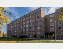 Fünfgeschossiges, rechteckiges Wohngebäude aus Betonplatten mit regelmäßiger Anordnung von Fenstern. Im Vordergrund ein Gehweg, ein Rasen mit Herbstlaub sowie Bäume; rechts ein offener Durchgang, der zu einem Hinterhof führt.