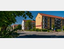 Mehrstöckige Wohnhäuser mit gelber und weißer Fassade, Balkonen und roten Dachziegeln stehen an einer Kreuzung. Im Vordergrund sind Bäume, Büsche und ein Verkehrsschild zu sehen, eingerahmt von klarem Himmel.