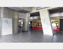 Ein überdachter S-Bahnhof mit einem gelb-roten Zug, der auf einem Gleis steht. Betonpfeiler mit Graffiti und eine metallene Wand mit einer Tür prägen die moderne, funktionale Architektur.