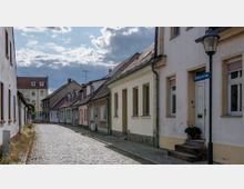 Gepflasterte Straße in einem historischen Wohnviertel mit kleinen Reihenhäusern und geneigten Ziegeldächern; auf der rechten Seite erkennbar ein Laternenpfahl und eine Hausnummer an der Tür. Im Hintergrund ein größeres Gebäude mit rotem Dach und mehreren Fenstern.