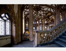 Historisches Gebäude mit gotisch inspiriertem Innenraum, geprägt durch hohe Spitzbögen, detaillierte Steinverzierungen und ein komplexes Skelettgewölbe; im Vordergrund ein Treppenaufgang mit kunstvoll durchbrochenem Geländer.