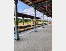 Bahnhof mit überdachtem Bahnsteig aus Holz und Metallstützen, gepflastertem Boden und Schienen im Vordergrund. Im Hintergrund sind weitere Bahnsteige, ein kleines Gebäude sowie grüne Vegetation bei sonnigem Wetter sichtbar.