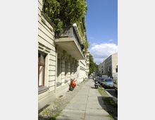 Straßenszene in einem Wohnviertel mit mehrstöckigen Gebäuden und Balkonen, die teilweise mit Pflanzen bewachsen sind. Entlang der Straße stehen Autos und Motorroller, und der Gehweg aus Betonplatten ist teilweise uneben.