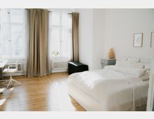 Helles Schlafzimmer mit Holzdielenboden, weiß gestrichenen Wänden und großen Fenstern, die von beigefarbenen Vorhängen eingerahmt sind. Ein Bett mit weißer Bettwäsche steht neben einem kleinen grauen Nachttisch, auf dem eine geflochtene Lampe steht, und neben dem Fenster befindet sich ein schwarzer Truhe.