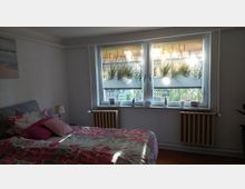 Schlafzimmer mit Doppelbett und floraler Bettwäsche, gegenüber ein breites Doppelfenster mit halbtransparenten Rollos im Dünenmotiv und kleiner Fensterbankdekoration. Unter dem Fenster stehen zwei Heizkörper; der Raum hat helle Wände und Holzboden.